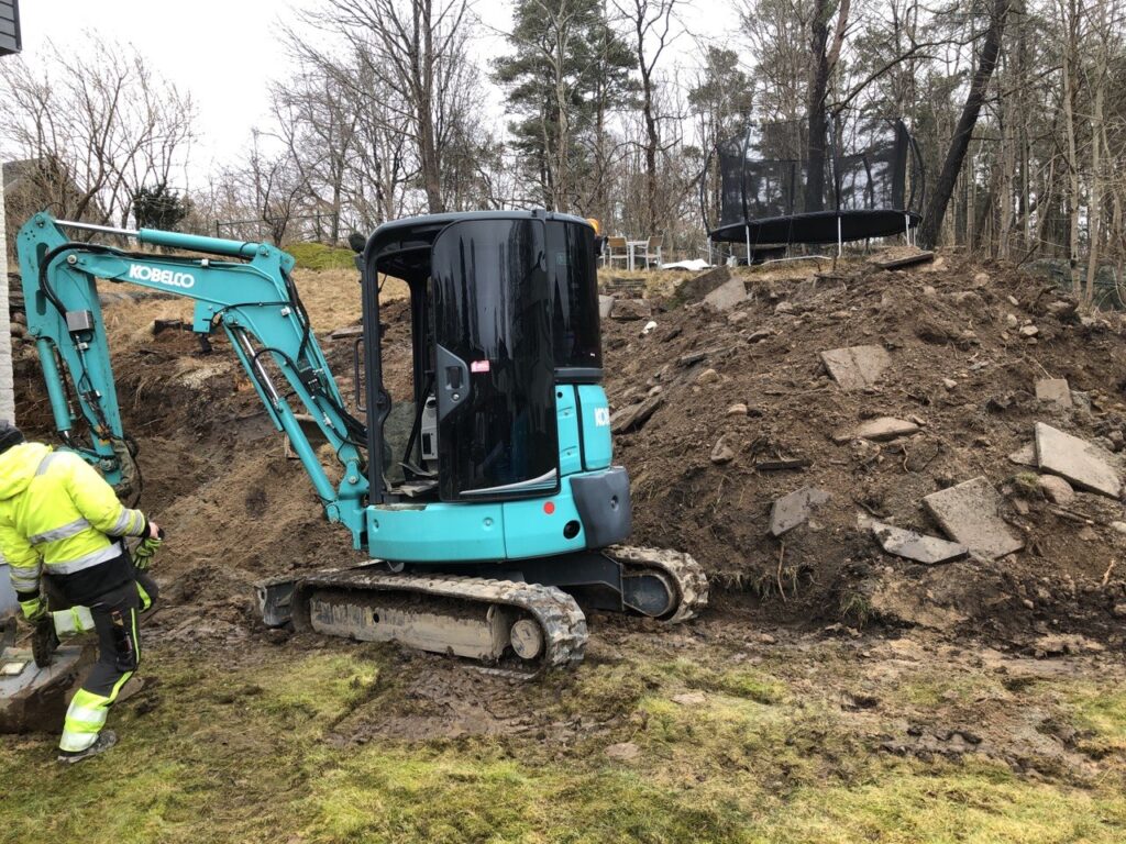 Grävmaskin vid schaktning runt husgrund under dräneringsarbete i Trollhättan.