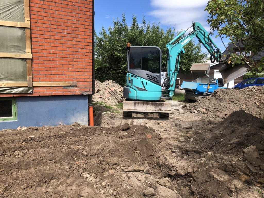 Minigrävare schaktar runt husgrunden inför dräneringsarbete i Vänersborg.